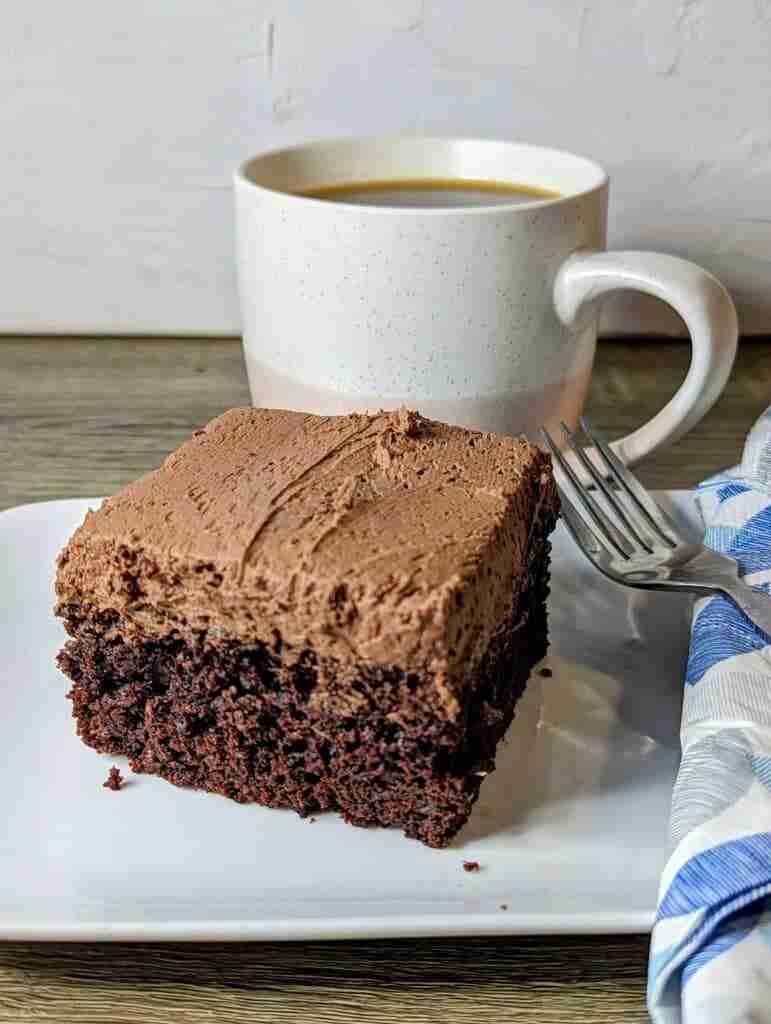 Air Fryer Chocolate Cake on a white plate with a fork and blue napkin. a cup of coffee in the background.