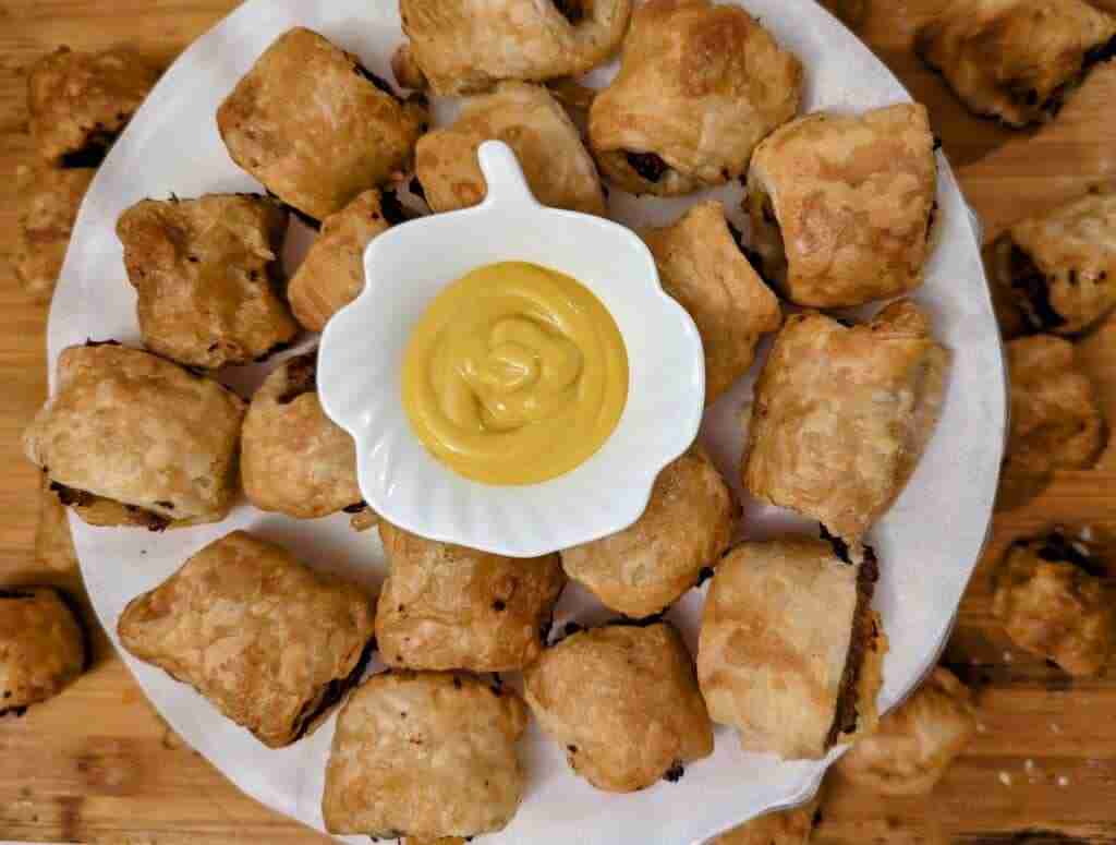 a white plate filled with homamde sausage rolls and a small bowl of mustard in the middle with more sausage rolls in the background