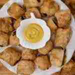 a white plate filled with homamde sausage rolls and a small bowl of mustard in the middle with more sausage rolls in the background