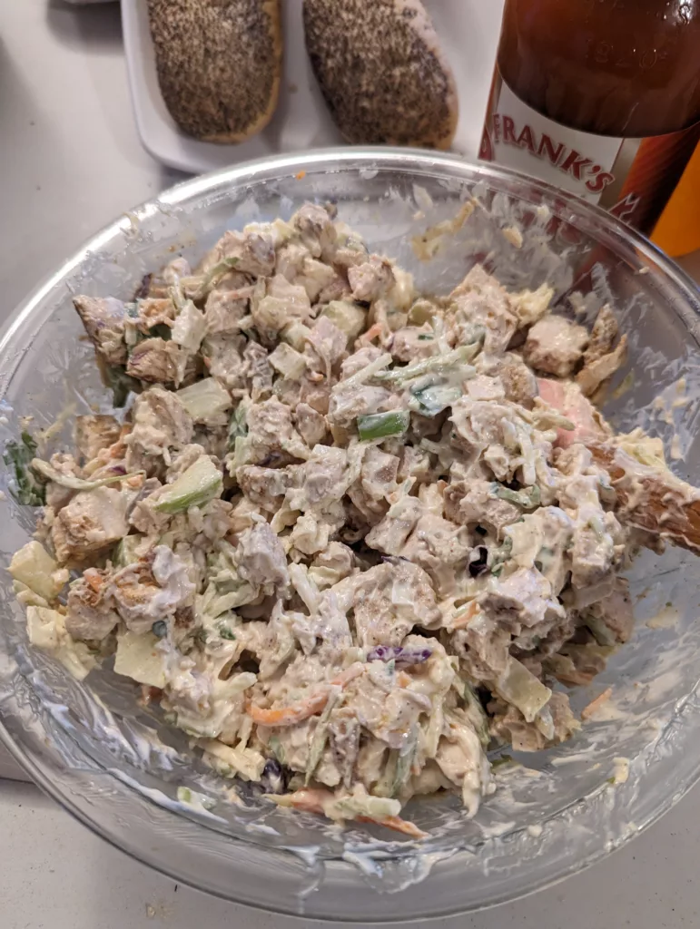 Chicken salad in a bowl with poppy seed buns and hot sauce in the background