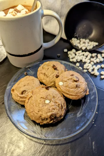 hot chocolate cookies on a plate with a cup of hot chocolate with marshmallows and a bowl of mini marshmallows spilled in the backround