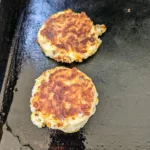 two sausage cheddar biscuits cooking on a flat top griddle