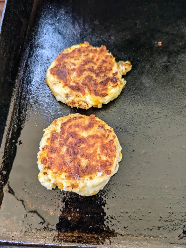 two sausage cheddar biscuits cooking on a flat top griddle