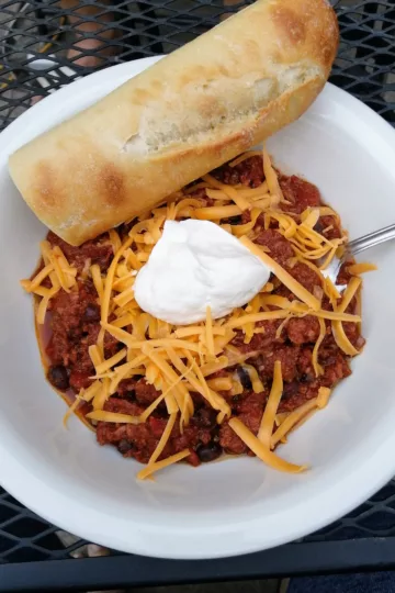 Bowl of chili topped with a dollop of sour cream and shredded cheese with a half baguette resting on the side of the bowl
