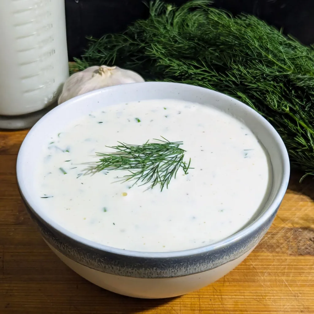 A bowl of Creamy Buttermilk Ranch Salad Dressing with fresh dill on top. There is a cup of buttermilk and more fresh dill behind the bowl.