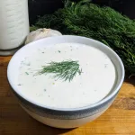 A bowl of buttermilk ranch sauce with fresh dill on the top. There is a cup of buttermilk and more fresh dill behind the bowl.