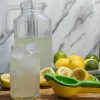 A glass of homemade lemonade and a glass pitcher of lemonade. There is also a green and yellow juicer and lemons and limes beside the lemonade.