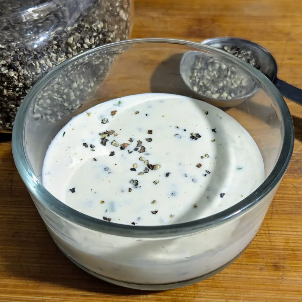 A clear glass bowl of Creamy Peppercorn Ranch Sauce with a jar of peppercorns and a spoon of peppercorns in behind the bowl.
