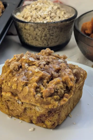 Pumpkin spice oatmeal muffin on a plate with a bowl of oats and a bowl of pumpkin puree in the background.