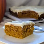 A piece of pumpkin spice cake with cream cheese icing on a white plate with the rest of the cake behind it. There is a pumpkin barely showing to the left.