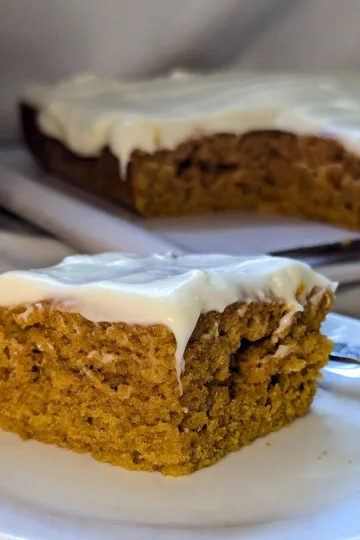 A piece of pumpkin spice cake with cream cheese icing on a white plate with the rest of the cake behind it. There is a pumpkin barely showing to the left.