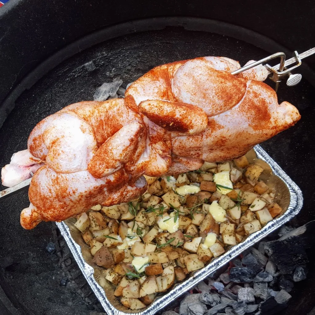 Two whole chicken on a BBQ rotisserie over a pan of potatoes.