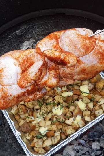 Two whole chicken on a BBQ rotisserie over a pan of potatoes.