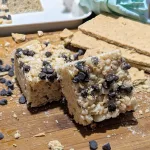 Two S'mores Rice Krispies on a cutting board with graham crackers off to the right side and chocolate chips mixed with graham crumbs.