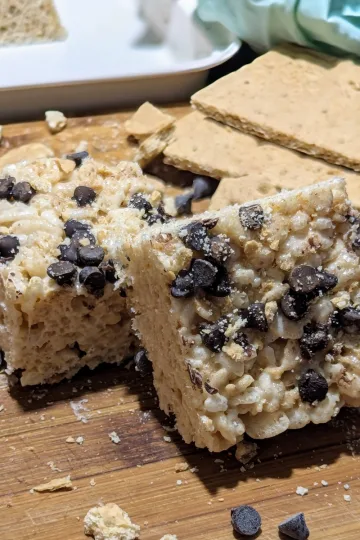 Two S'mores Rice Krispies on a cutting board with graham crackers off to the right side and chocolate chips mixed with graham crumbs.