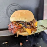 Meatloaf burger with mushroom relish on a spatula held over a Blackstone griddle.