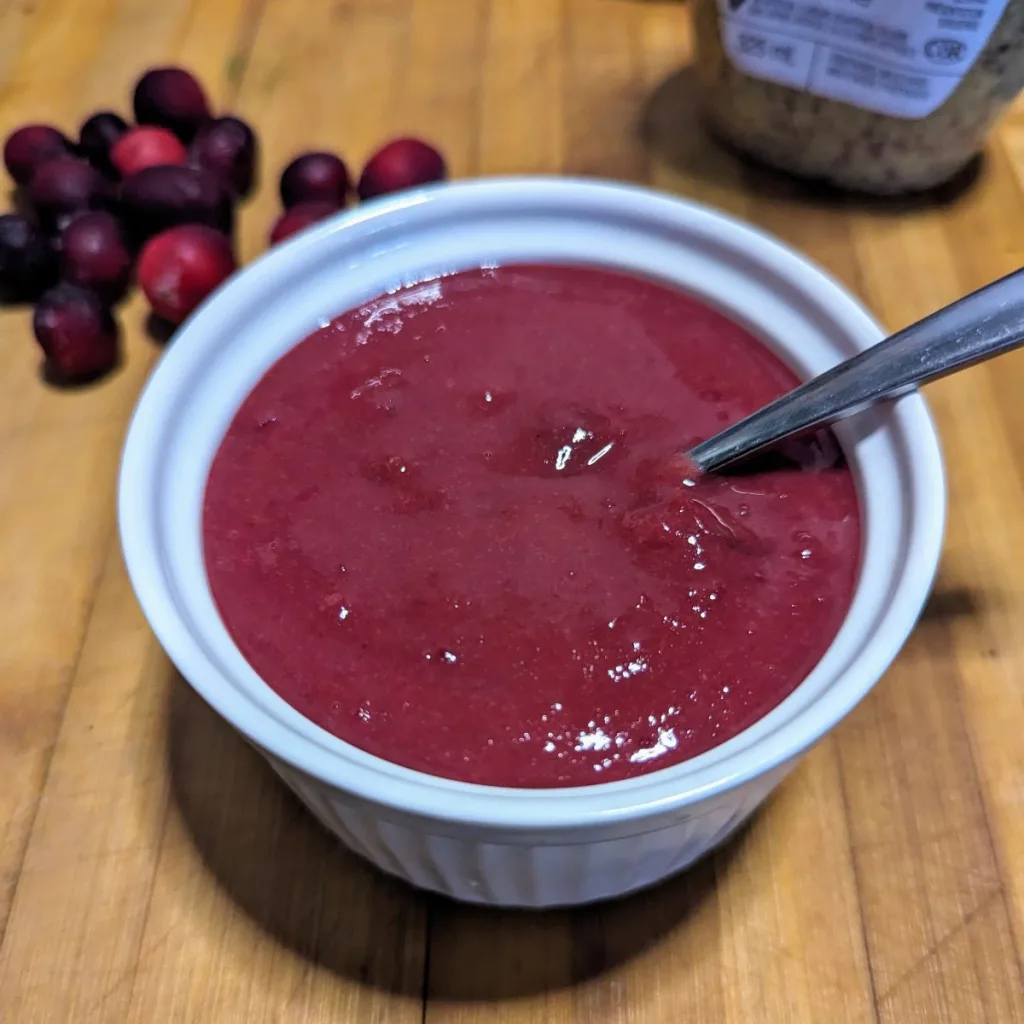 A small bowl of Cranberry Mustard Dipping Sauce with a small spoon in it. There are fresh cranberries off to to left.