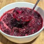 A white bowl filled with homemade cranberry sauce with a spoonful of cranberry sauce held over the bowl.