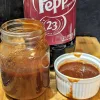 A white ramekin of Dr Pepper rib sauce with a jar half filled with sauce on the left and a bottle of Dr Pepper behind it.