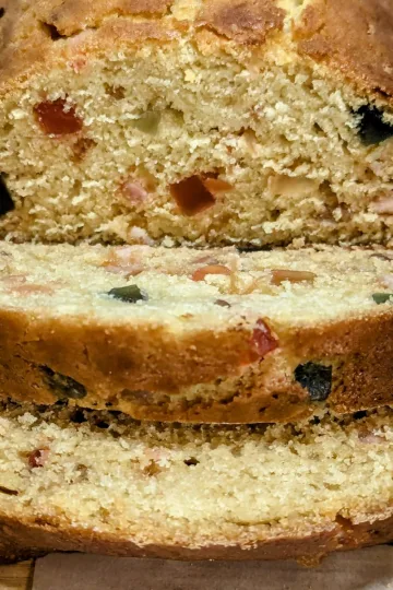 A fruitcake loaf on a cutting board. There are two slices lying in front of the cut loaf revealing the candied fruit in the loaf.