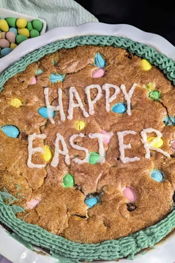 A giant mini egg cookie decorated with green icing around the edge and Happy Easter written in capital letters in white icing in the center of the cookie.
