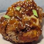 Fried chicken coated in gochujang sauce with sesame seeds and green onions sprinkled over.