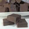 Three squares of homemade fudge placed in a pyramid form with a plate filled with a pile of squares of fudge behind it.
