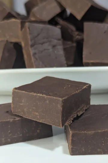 Three squares of homemade fudge placed in a pyramid form with a plate filled with a pile of squares of fudge behind it.