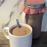 A white mug with hot chocolate and a spoon in it A mason jar of hot chocolate mix in the background with a red and white fabric lid.