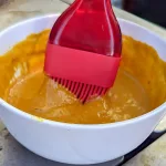 A bowl of sriracha mustard sauce with a red basting brush dipped in it.