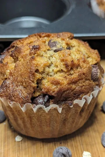 Chocolate chip oatmeal muffin with spilled chocolate chips around it and a muffin pan behind it.
