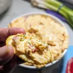 A cracker with pimento cheese being help over the bowl of pimento cheese.