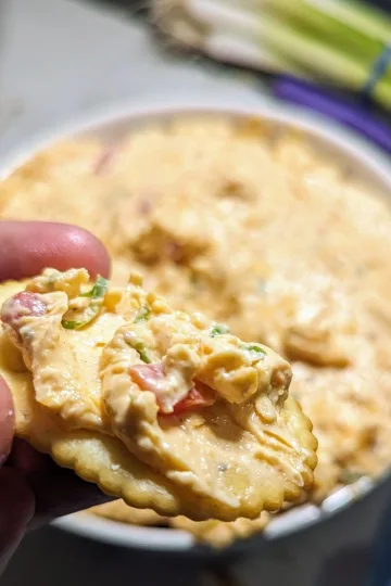 A cracker with pimento cheese being help over the bowl of pimento cheese.