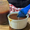 A small white bowl of pineapple BBQ sauce with a blue basting brush being dipped in it. There is a jar of sauce on the left side and a red and white checkered napkin lying behind.