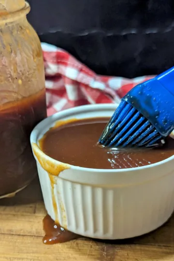 A small white bowl of pineapple BBQ sauce with a blue basting brush being dipped in it. There is a jar of sauce on the left side and a red and white checkered napkin lying behind.
