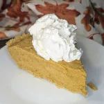 A slice of pumpkin chiffon pie topped with whipped cream. There is a cloth with fall leaves in the background.