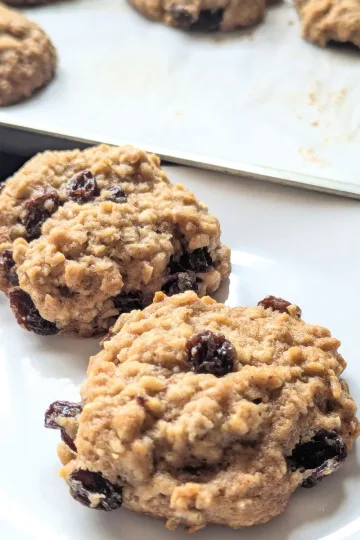 Two rum raisin oatmeal cookies on a plate with a tray of cookies in the back.