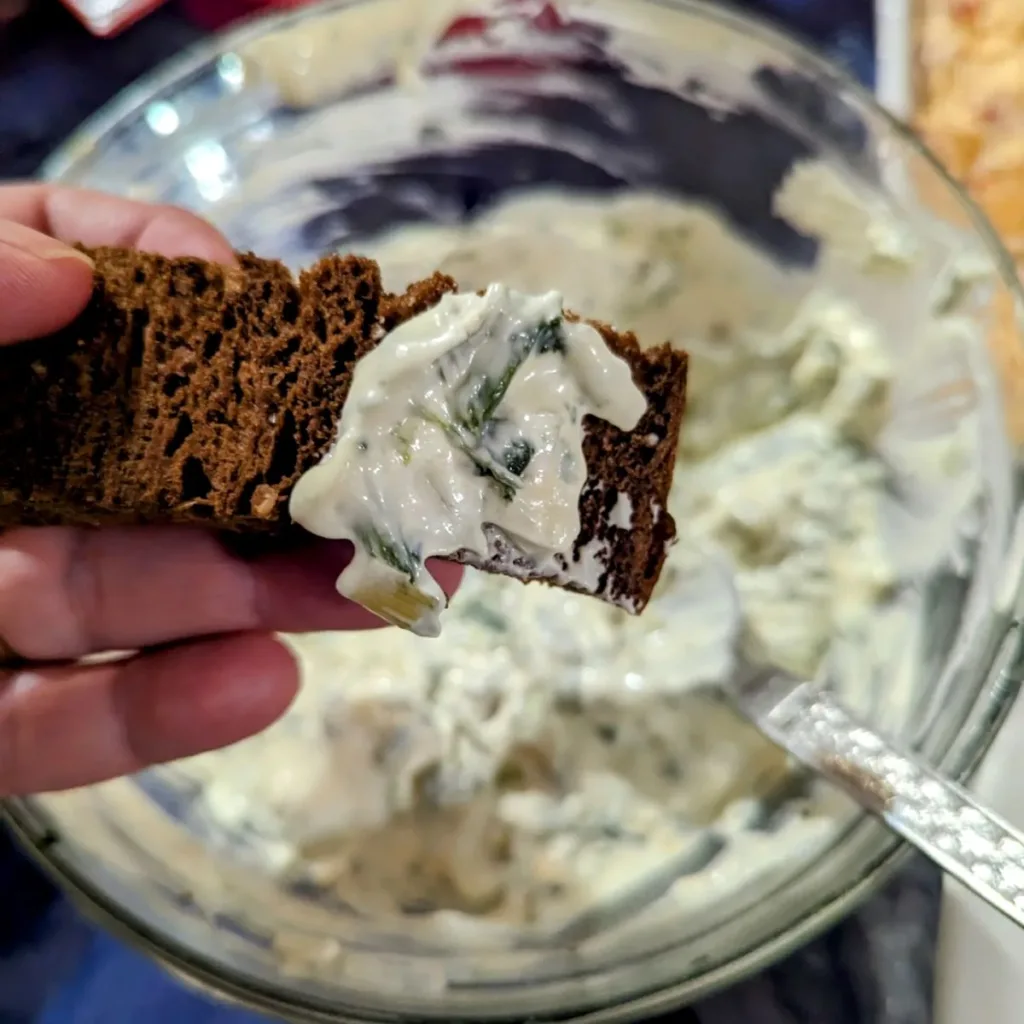 A small piece of Pumpernickel bread with Easy No Bake Spinach Dip on it being held over a bowl of spinach dip.