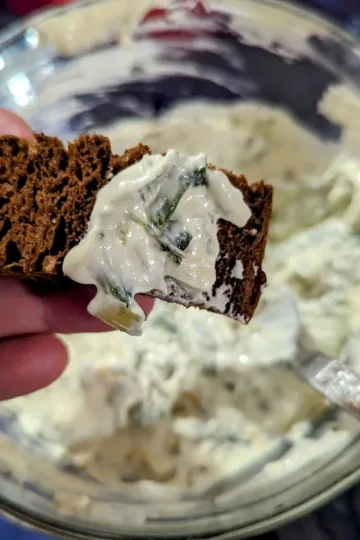 A small piece of Pumpernickel bread with spinach dip on it being held over a bowl of spinach dip.