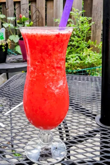 Strawberry Malibu Cocktail in a hurricane glass on a patio table. There are plants in the background.