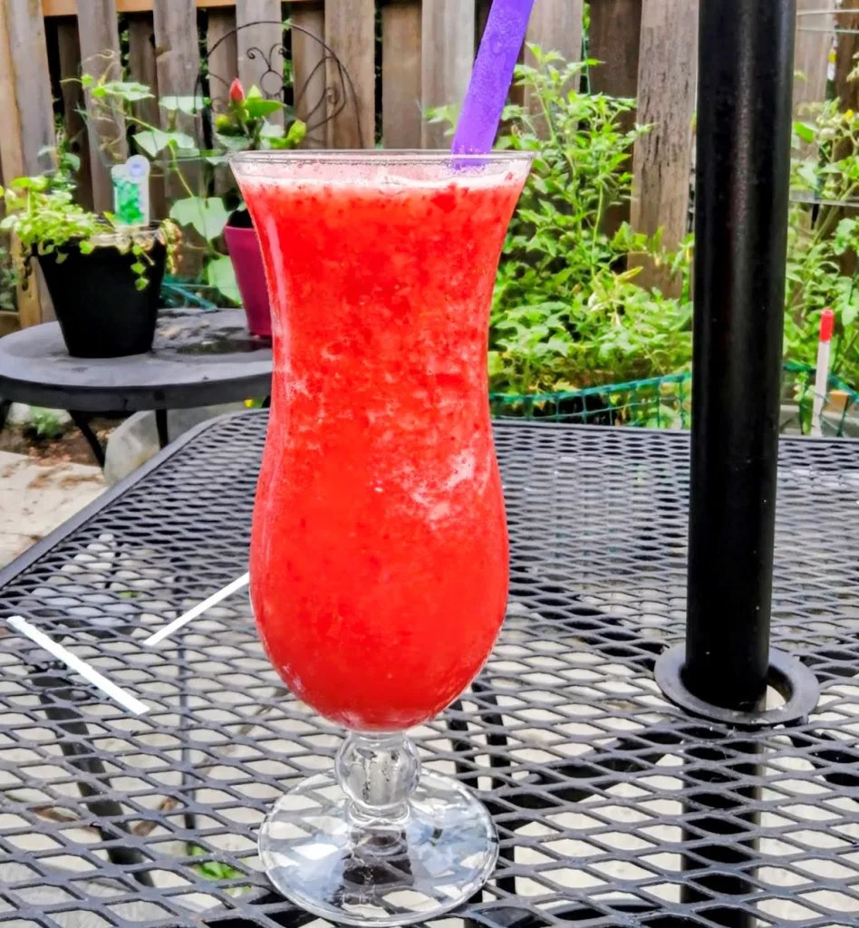Strawberry Malibu Slush in a hurricane glass on a patio table. There are plants in the background.