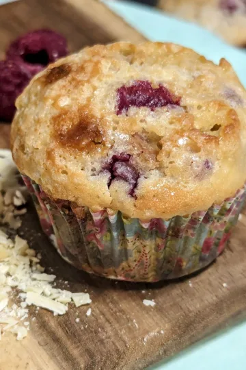 White chocolate raspberry muffin on a board with fresh raspberries and shaved white chocolate on the board.