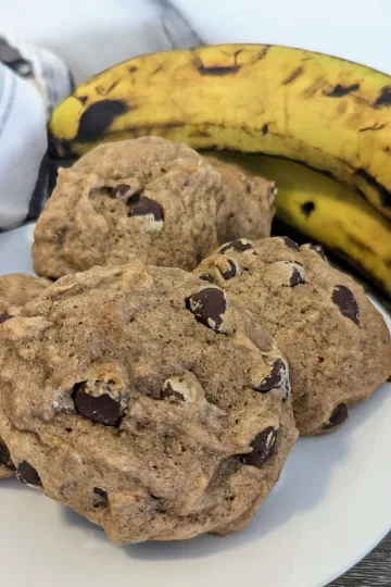 A plate of banana chocolate chip cookies with overripe bananas in the background.