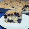 A slice of blueberry muffin cake on a plate with the rest of the cake in a pan in the background.