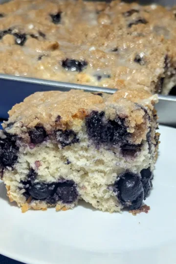 A slice of blueberry muffin cake on a plate with the rest of the cake in a pan in the background.