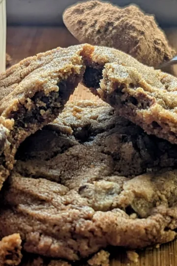 One Gingerbread chocolate chip cookie broken in half and laying on top of another cookie. There is a spoonful of gingerbread spice behind the cookies.
