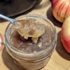 A jar of spiced apple jam with a spoonful of jam held over top with two apples barely visible off to the right side.
