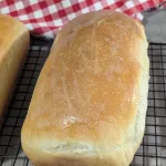 A loaf of homemade sandwich bread on a wire rack with a red and white towel in the background.