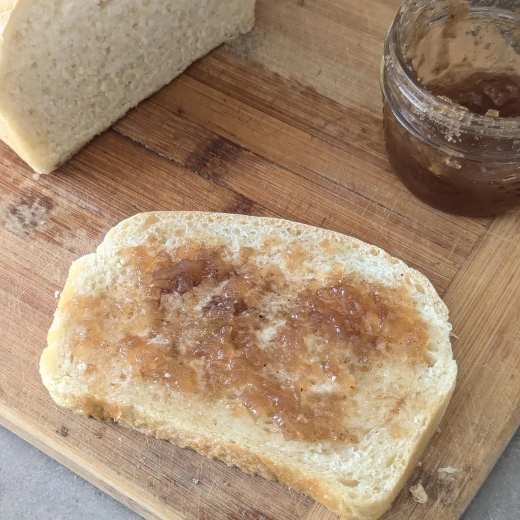 A slice of homemade bread with apple jam on it. There is a jar of jam in the background.