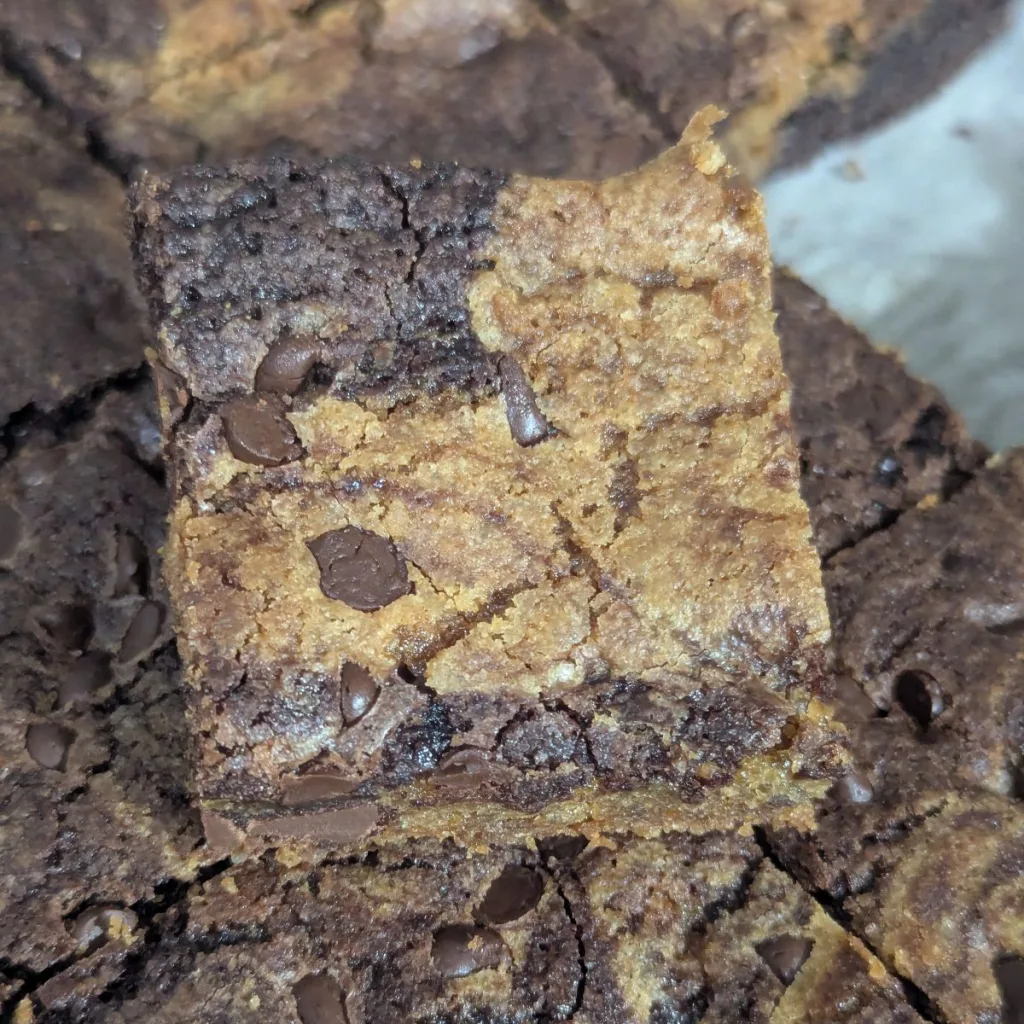 A cut square of brownie chocolate chip cookie bars sitting on top of the rest of the pan of bars.